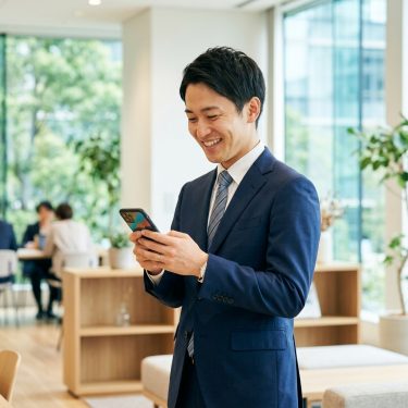 スマホで出会いアプリを探す笑顔のサラリーマン。昼間の明るい室内背景